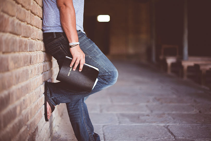 Man leaning against a brick wall holding a bible.