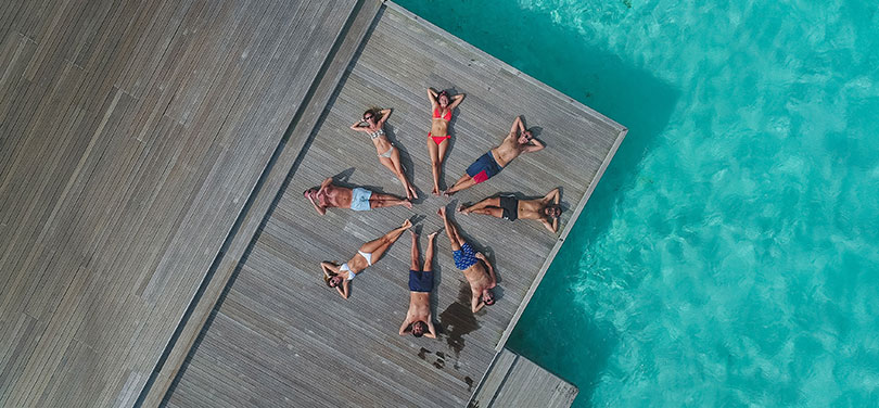 Young people sunbathe in a circle on a deck surrounded by crystal clear water. 