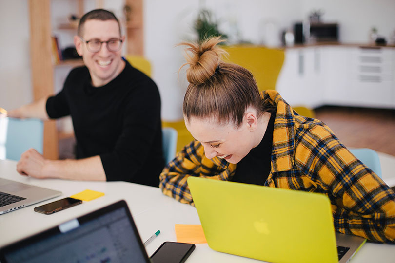 Woman laughing while sitting a desk next to a smiling man.