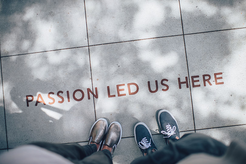 The words "passion led us here" are stamped on sidewalk and two people stand below it but only their shoes and legs are visible.