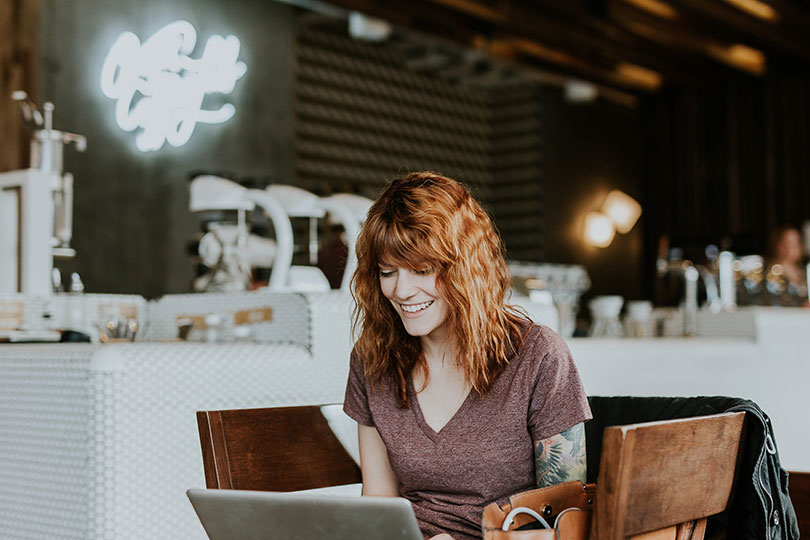 Woman sitting in a café looking at a laptop screen smiling. 
