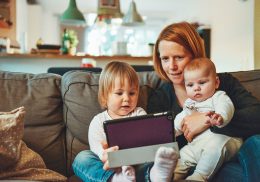 Mom sits on a couch with two babies and a tablet.
