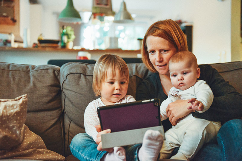 Mom sits on a couch with two babies and a tablet. 