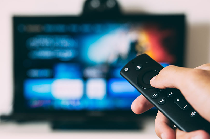 Hand holding a TV remote with screen in background. 