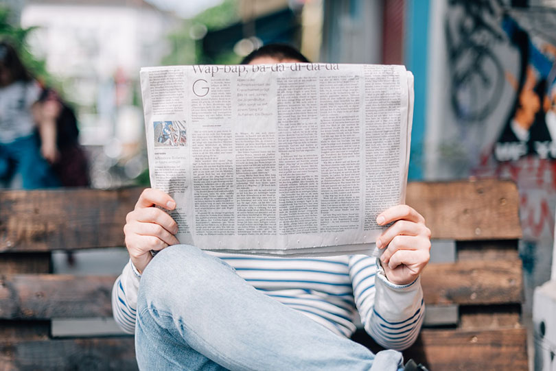 Man sitting on a bench reading a newspaper that covers his face.