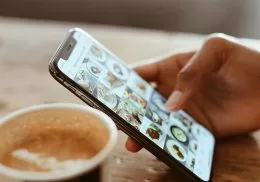 Close up image of hand scrolling through phone with a coffee next to it.